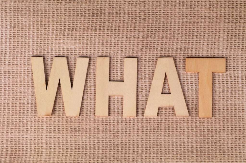 Wooden letters forming the word WHAT set on a textured burlap surface.