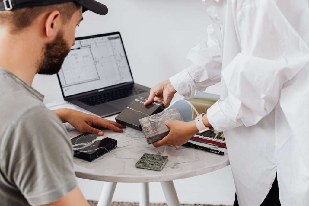 Two designers selecting marble samples indoors with laptop on table.