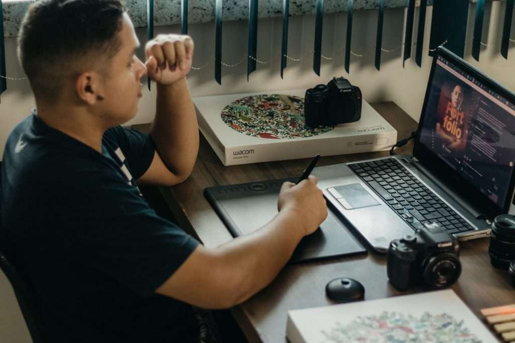 A photographer working at a creative setup with a laptop and Wacom tablet, focused on editing photos indoors.