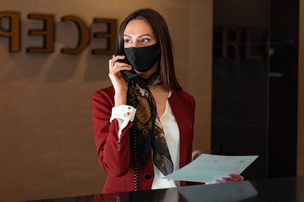 A female receptionist with a face mask answers a phone call, embodying professional service and safety.