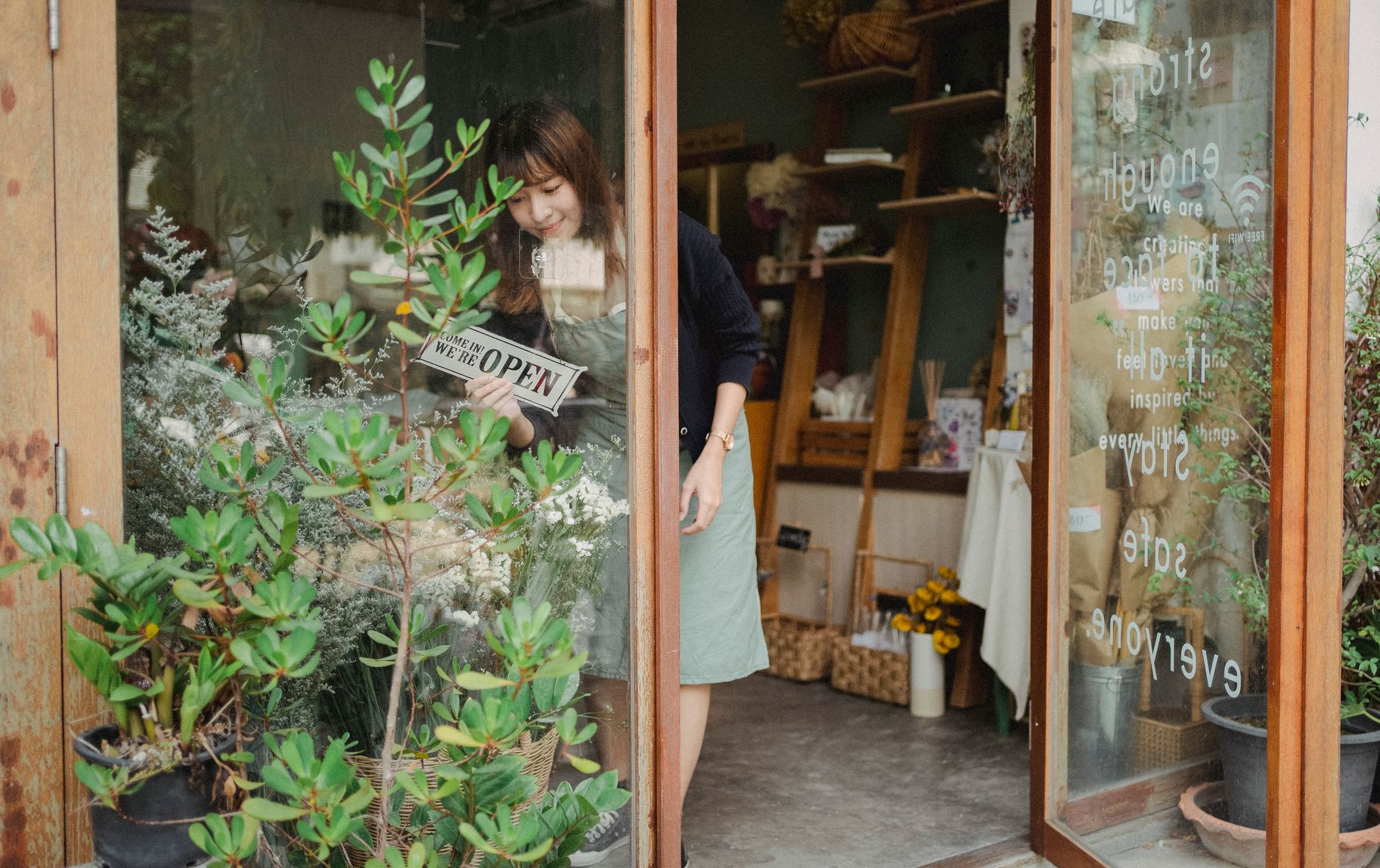 Positive young Asian female florist in apron standing near entrance and turning signboard on glass wall while working in m…